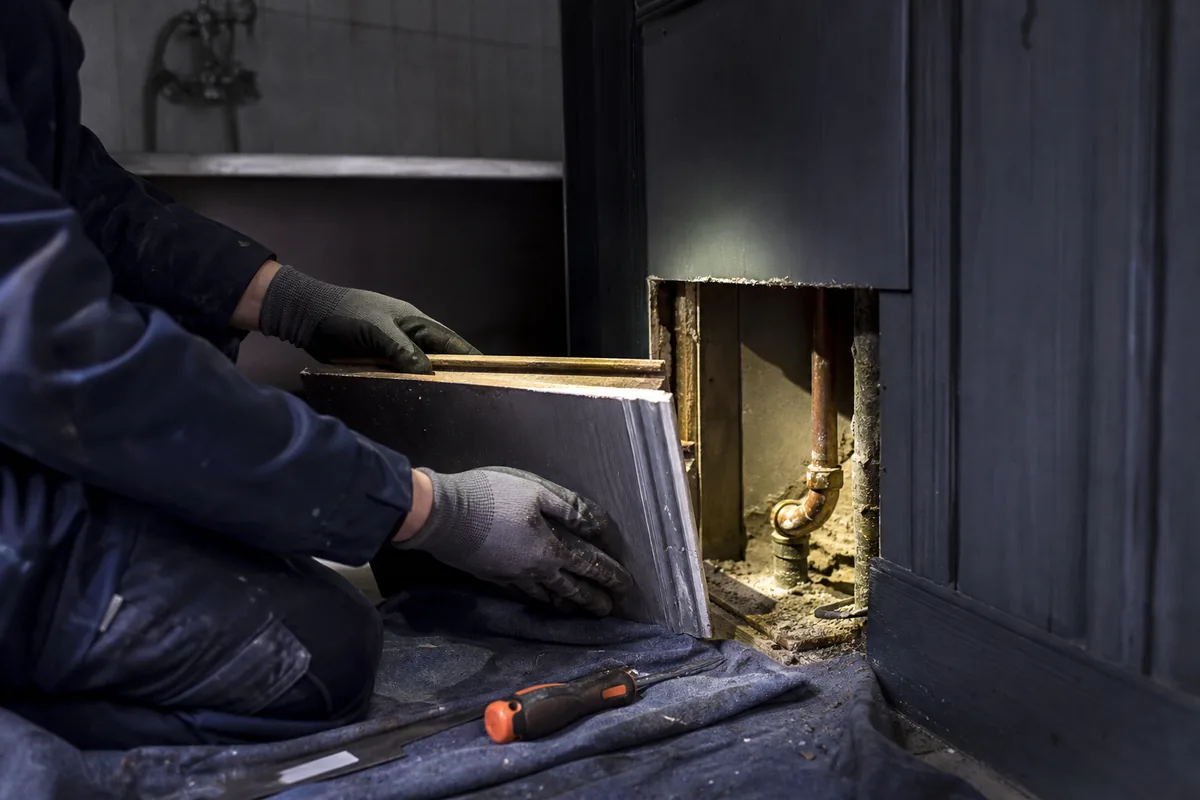 Mains gantées ouvrant un coffrage bois pour accéder à une canalisation encastrée dans une villa ancienne