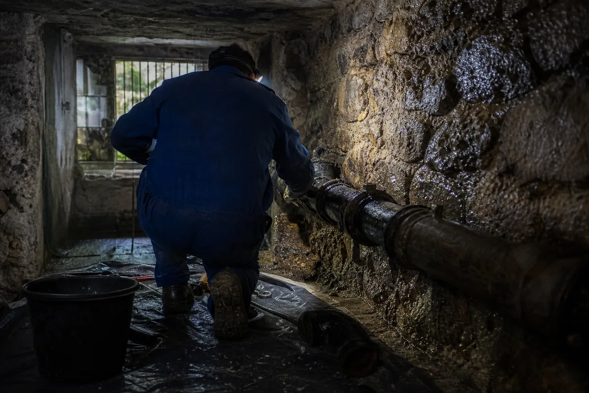 Plombier de dos éclairant un collecteur en fonte dans une cave semi-enterrée