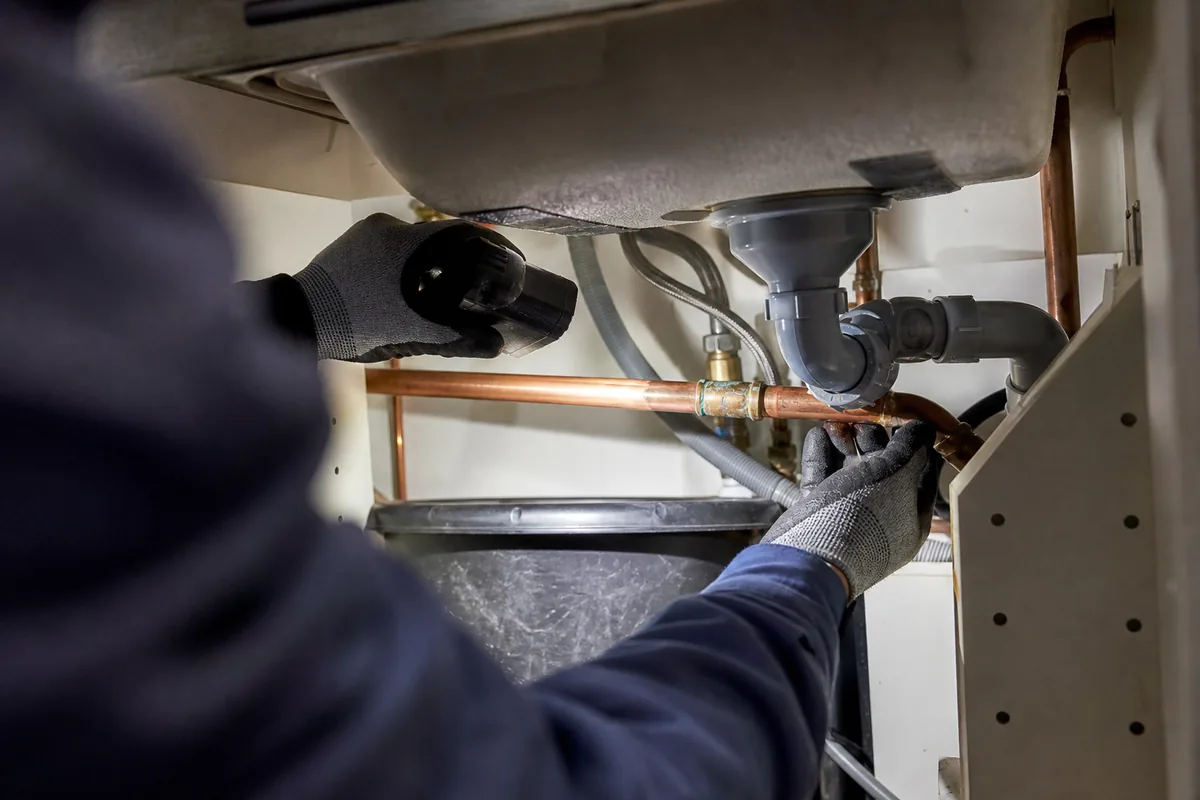 Mains gantées inspectant un raccord de cuivre corrodé sous un évier